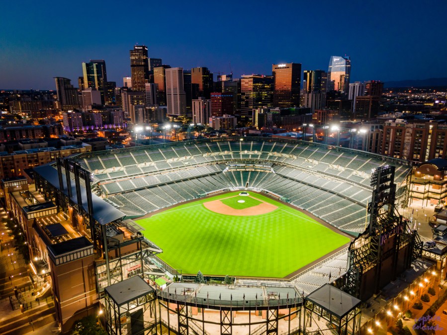 Aerial view of Coors Field at sunrise with baseball field lit up and skyline in the background