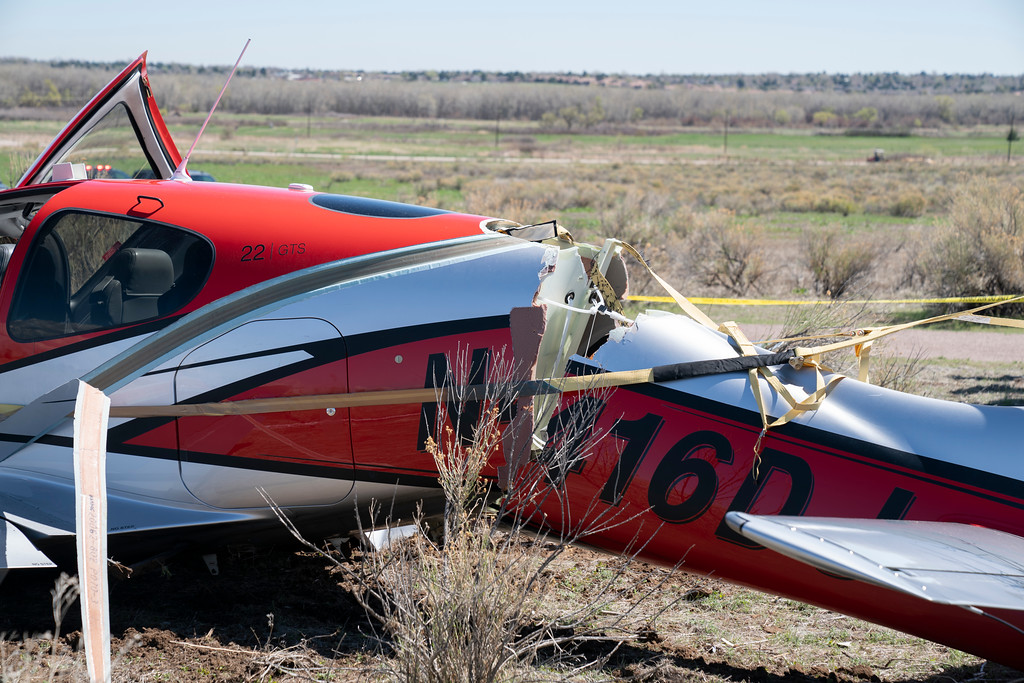 A small red and white airplane with a cracked fuselage and police tape surrounding it on a dry dirt field with blue sky on the horizon