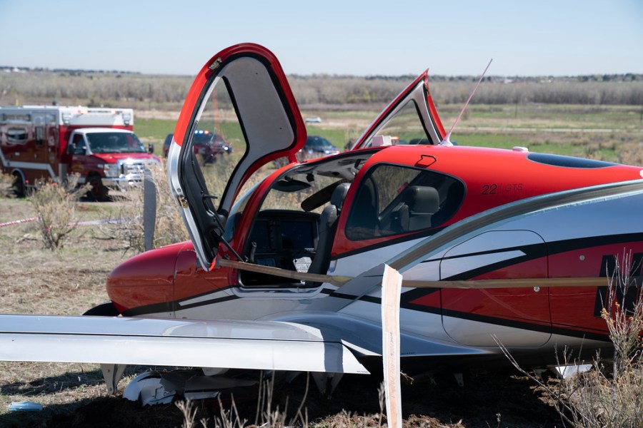 The side of a small red and white airplane with its two cockpit doors open and emergency vehicles in the background in a field with blue sky