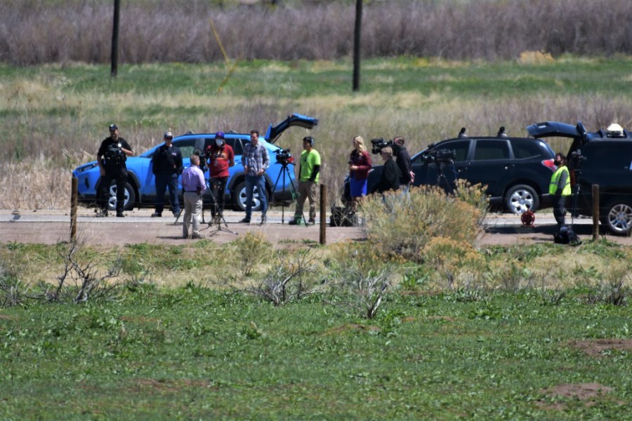 People lined up along the road with media cameras in front of two SUVs and surrounded by green grass fields