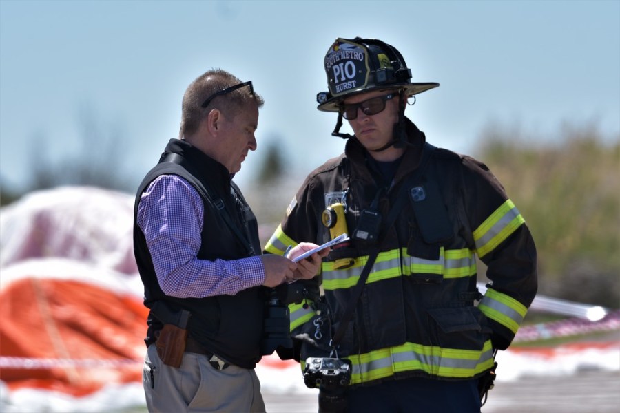 A closeup of a man in business attire talking to another man in firefighter attire with blue sky and the plane's parachute in the background