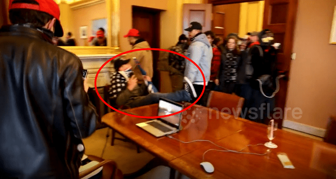 A man wrapped in an American flag sits at the head of a wooden conference table in a Capitol office and is surrounded by other people