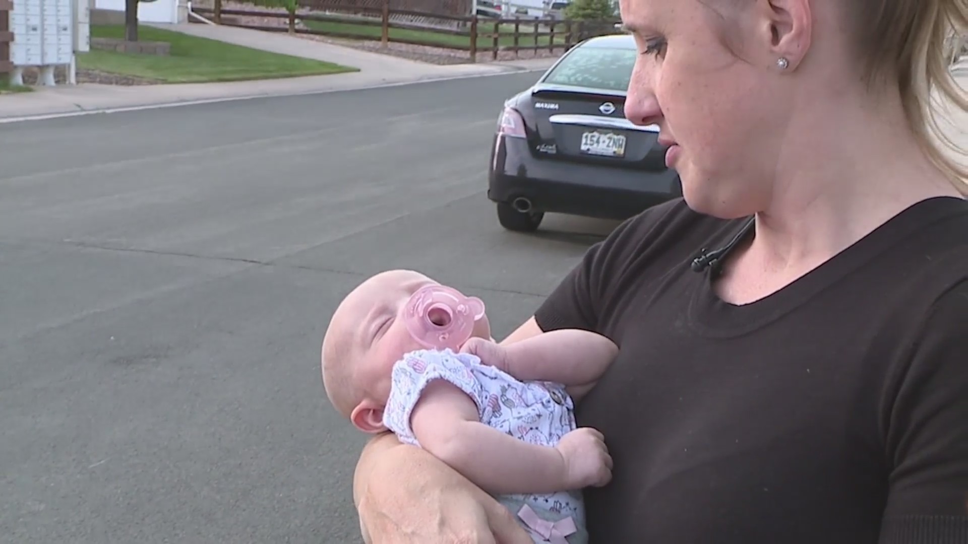 A woman holds a sleeping baby in her arms and looks down on her with roadway and a car in background