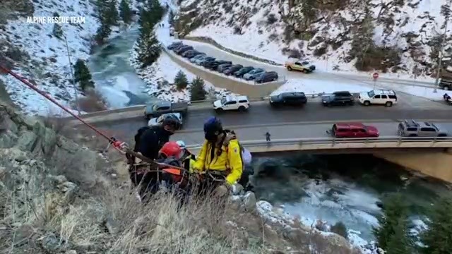 2 people carrying someone off a mountainside over a rushing creek near a parking lot