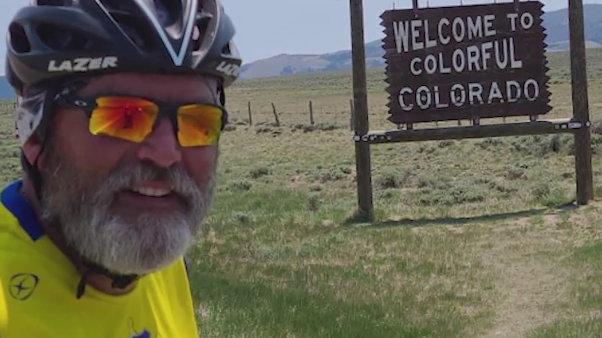 A close-up of a man with sunglasses and a helmet and a white beard and mustache with a field in the background with a "Welcome to Colorful Colorado" wooden sign and mountains in the distance