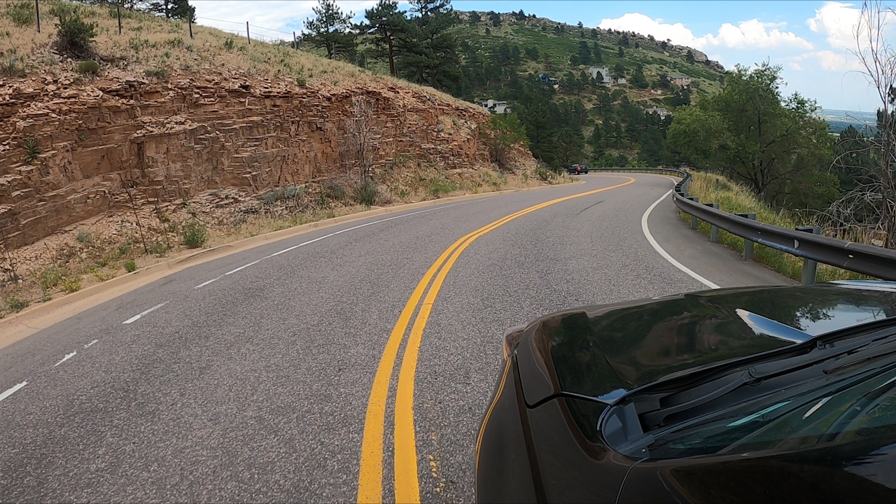 A paved road with double yellow center lines winds around the side of the foothills in daylight