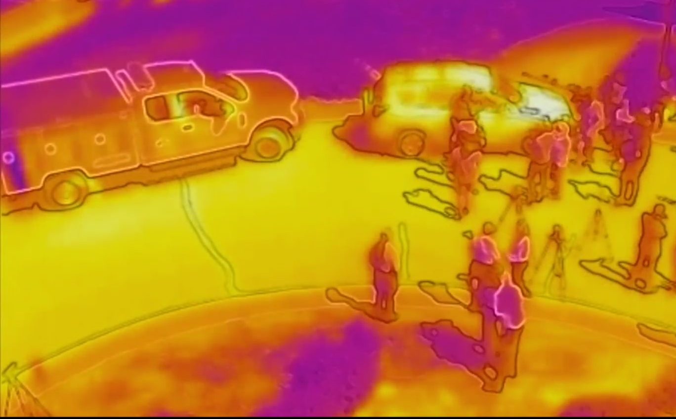 Infrared image of people on the street near two vehicles, image is colored pink, orange and yellow