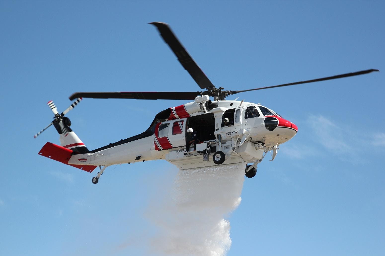 S70i Firehawk helicopter in flight with white substance spewing toward the ground from beneath it