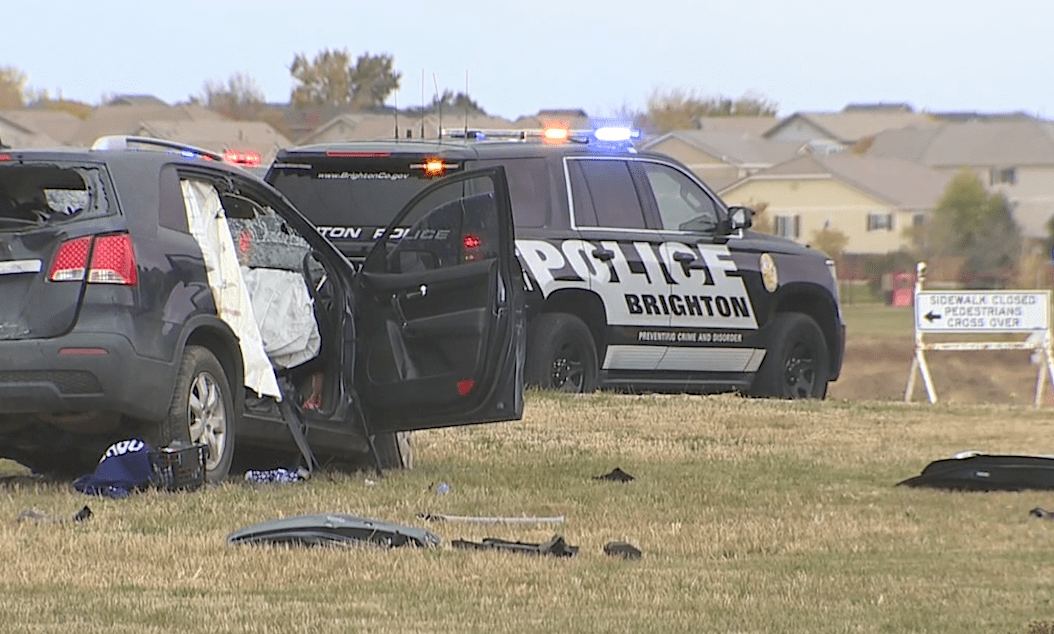 A damaged SUV with rear and front window busted out and parts strewn about next to Brighton police SUV in a field near homes