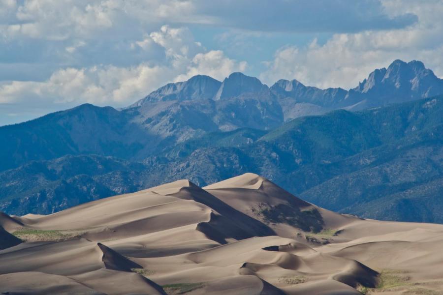 Sand dunes with mountains and a cloudy blue sky in the background