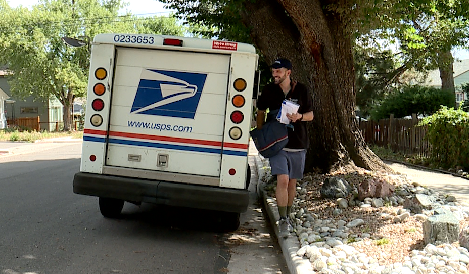 A mail carrier beside a U.S. Postal Service mail truck on a neighborhood road