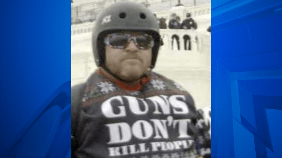 Man wearing helmet, sunglasses, shirt that says "GUNS DON'T KILL PEOPLE" with police on the Capitol balcony in the background