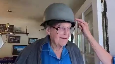 Older white man wearing black-rimmed glasses and an old military helmet looks off frame