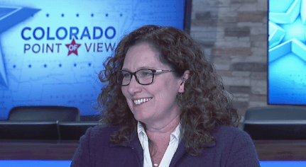 White woman with shoulder length curly hair and black-rimmed glasses smiles