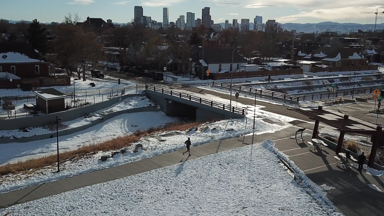 A man runs on a city trail with the Denver skyline in the background