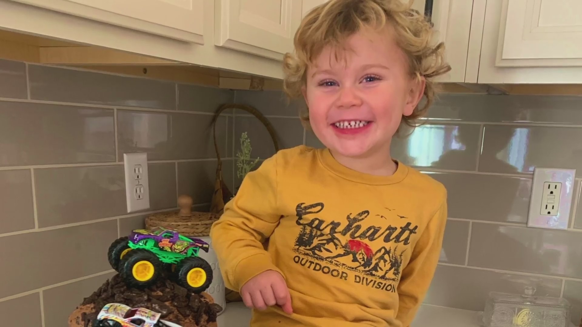 A little boy sitting on a kitchen counter next to a toy truck smiles for the camera