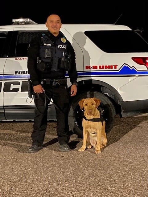 A police officer in uniform in front of a police SUV as he holds a K-9 dog on a leash