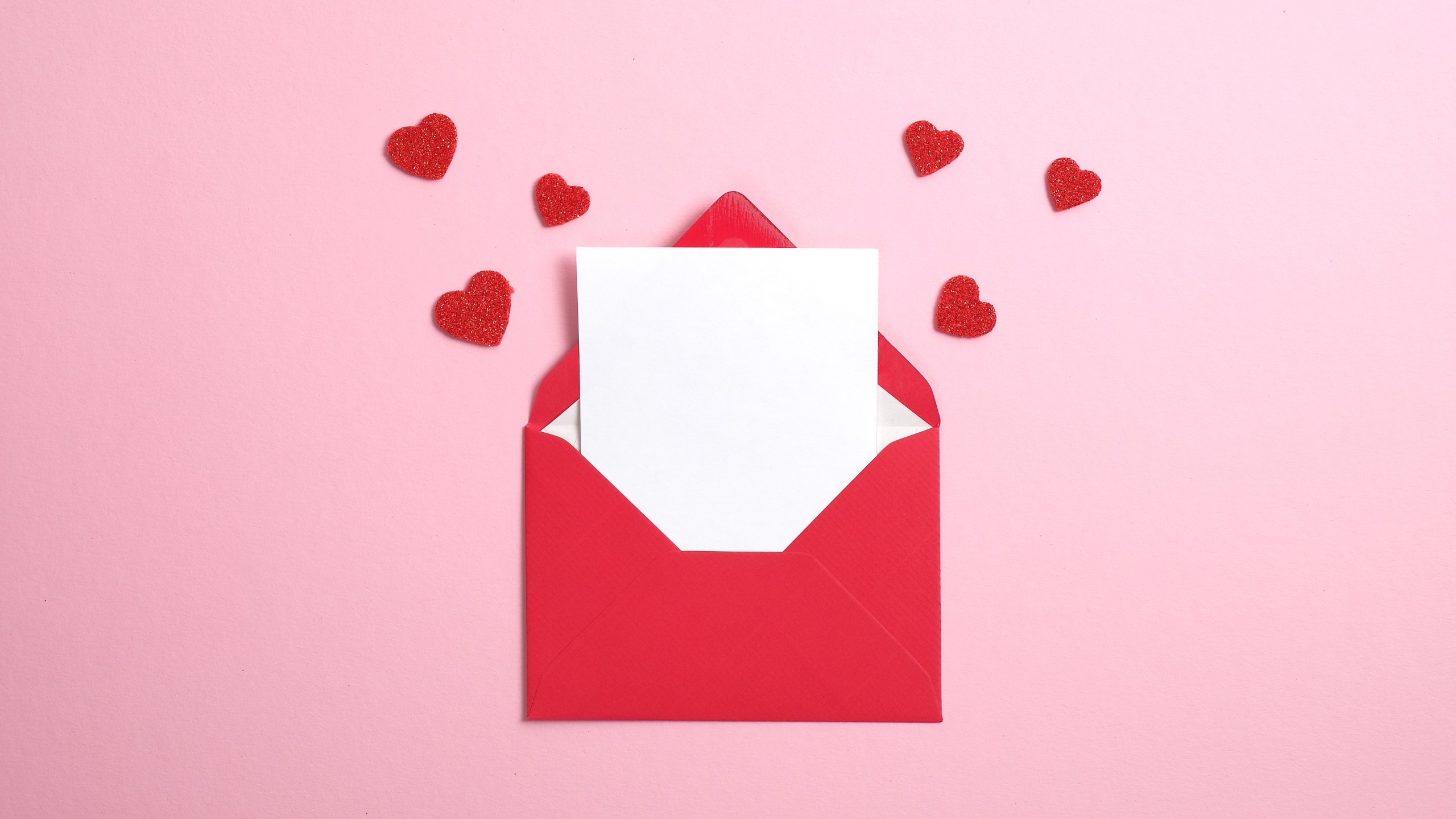 Red paper envelope with blank white note mockup inside and Valentines hearts on pink background. Flat lay, top view. Romantic love letter for Valentine's day.