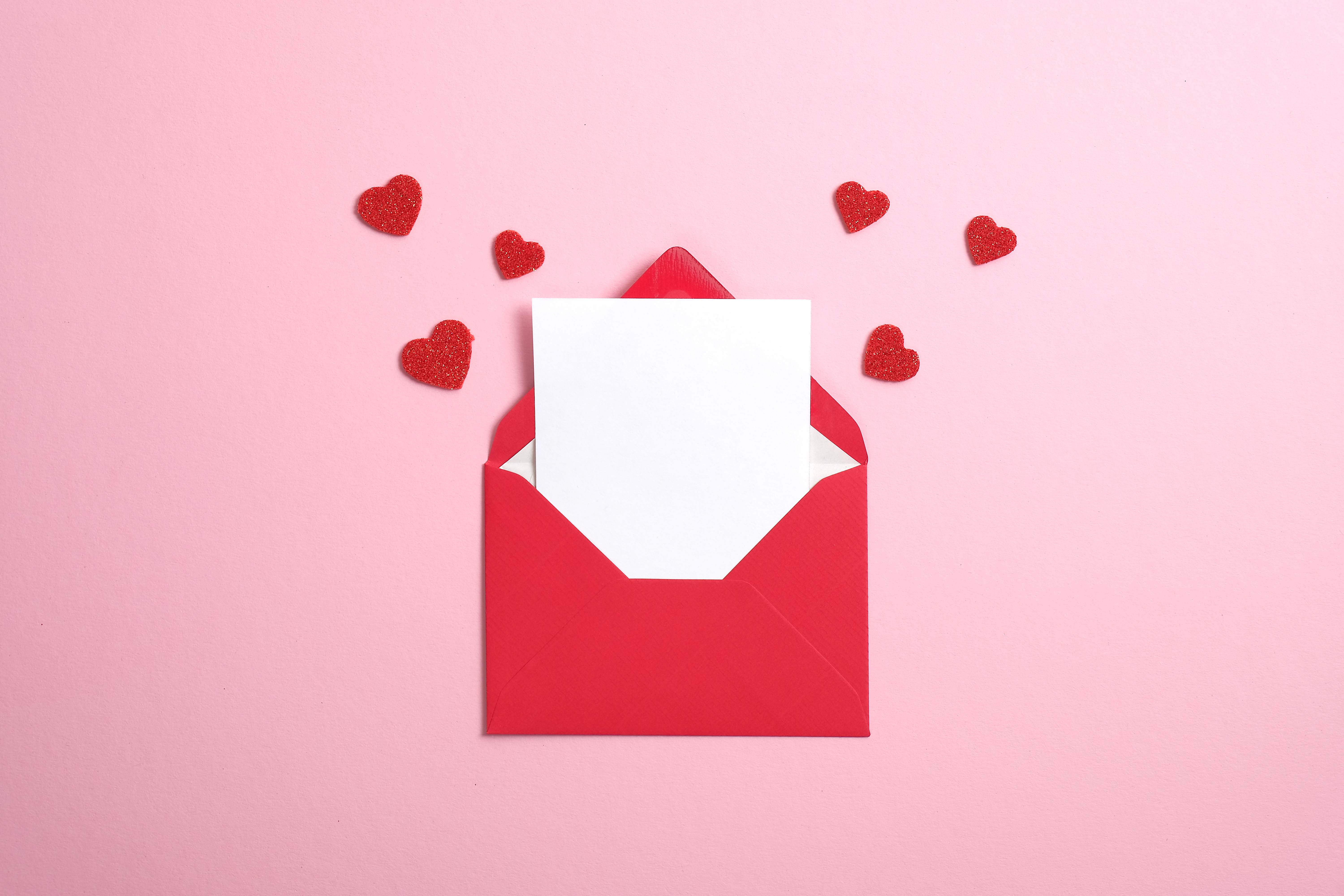 Red paper envelope with blank white note mockup inside and Valentines hearts on pink background. Flat lay, top view. Romantic love letter for Valentine's day.