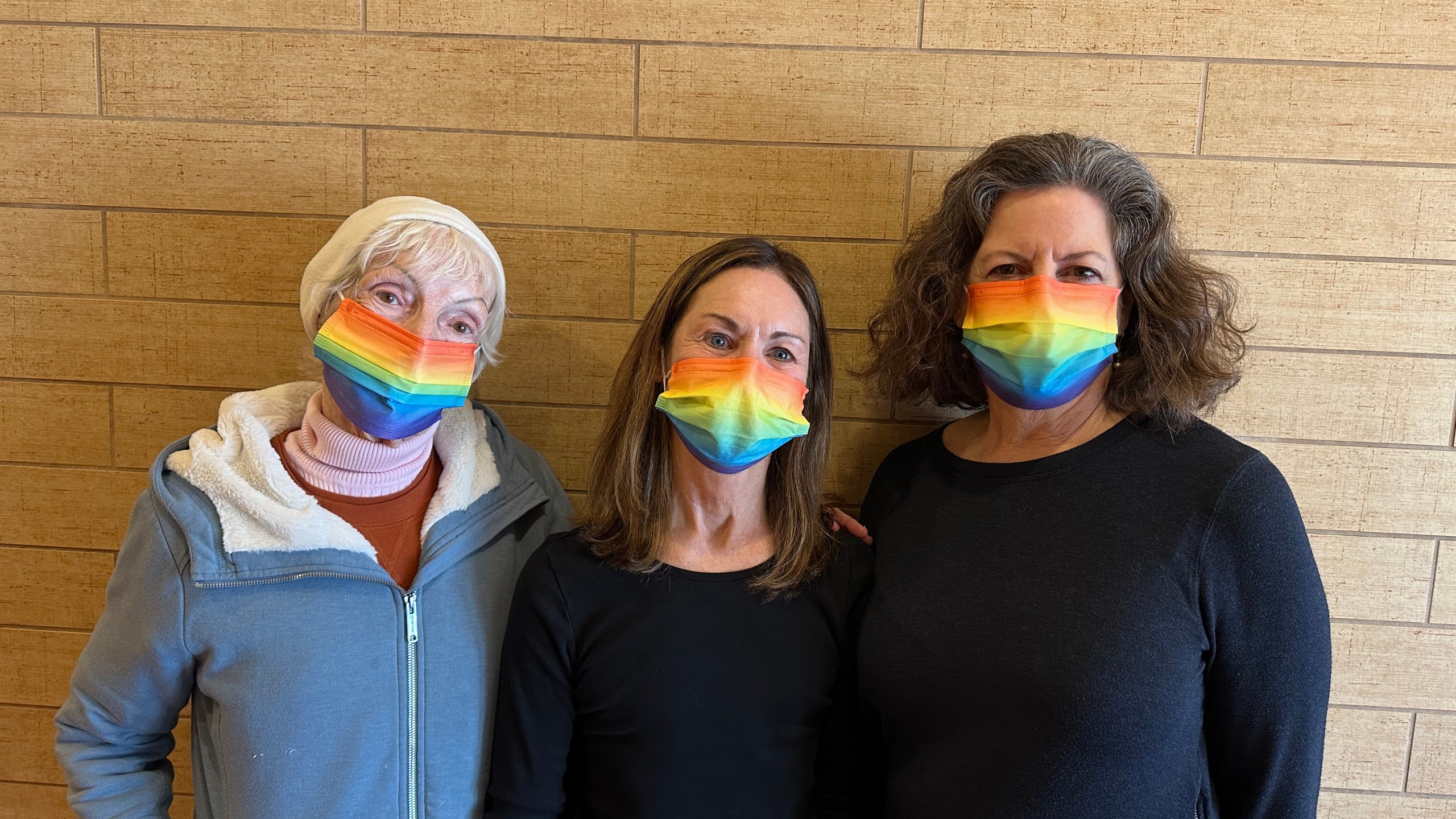 Three women stand side by side while wearing rainbow-colored face masks