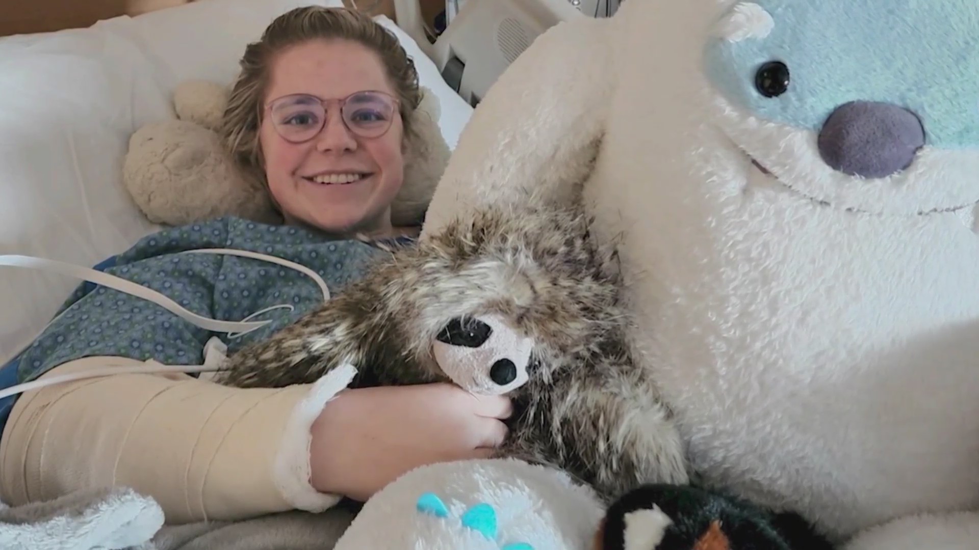 A woman smiling in a hospital bed with an arm cast and stuffed animals around her