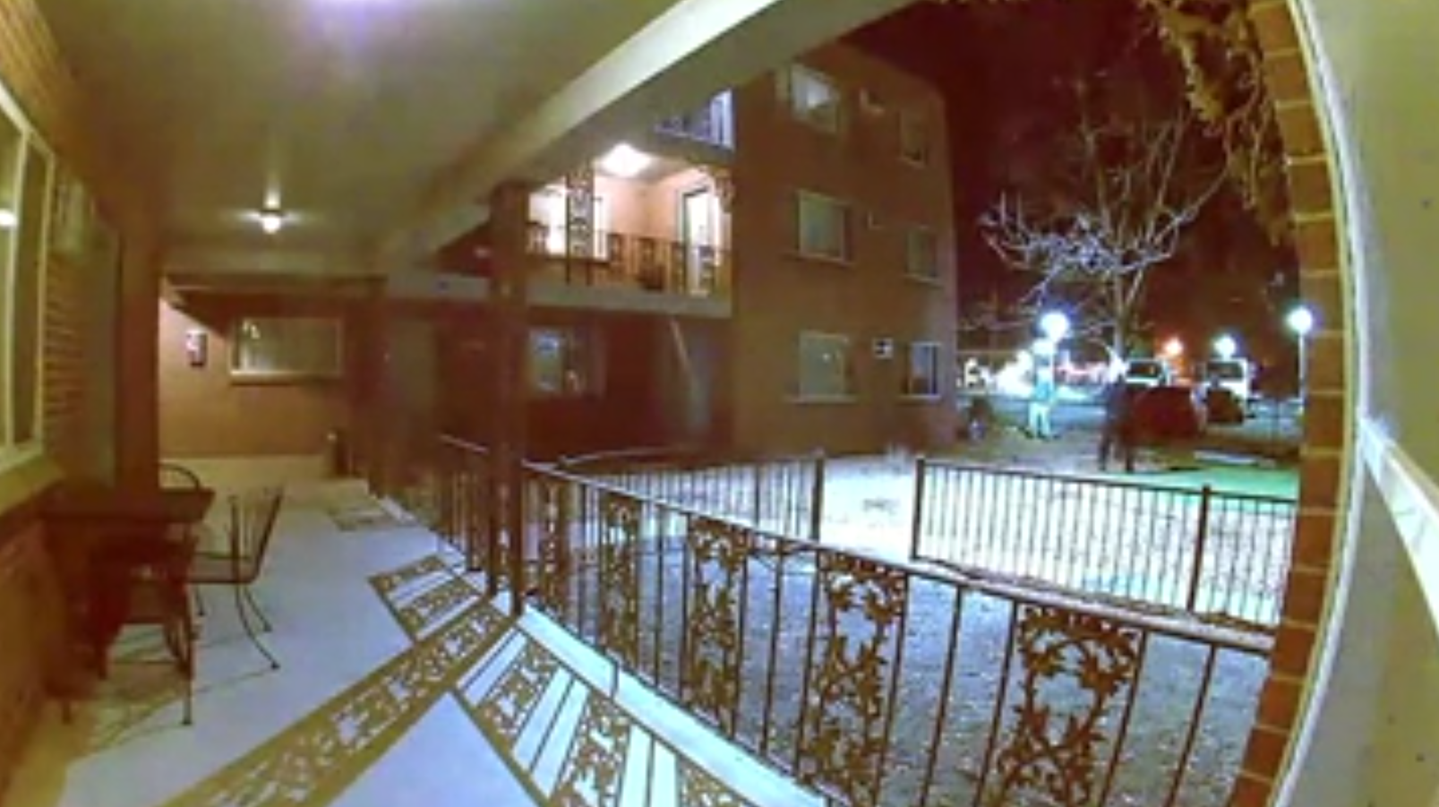 Screenshot of surveillance video showing an apartment courtyard and a police officer in the far corner facing a person who is lit with a flashlight