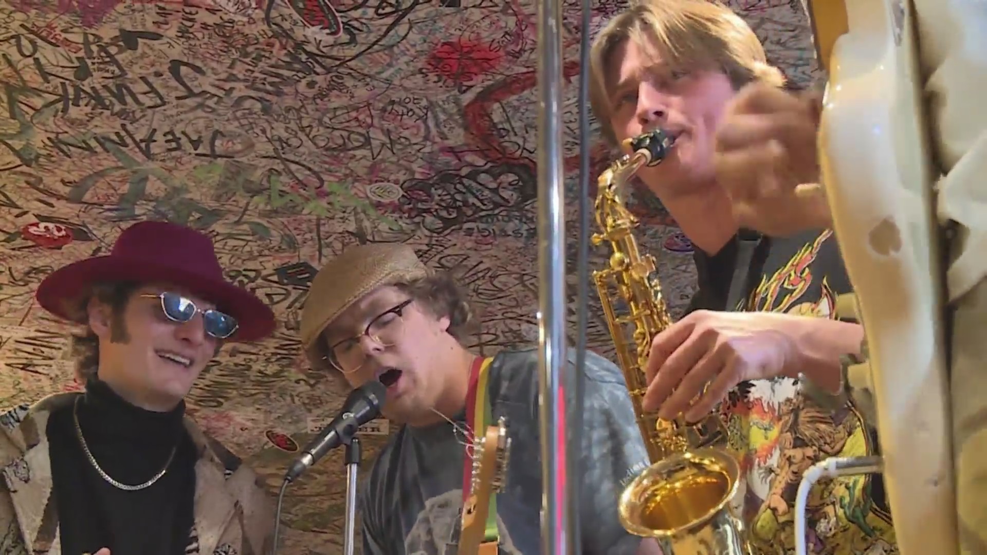 A singer plays guitar next to a blowing saxophinist and a man with a hat and funky sunglasses in front of a wall covered in writing