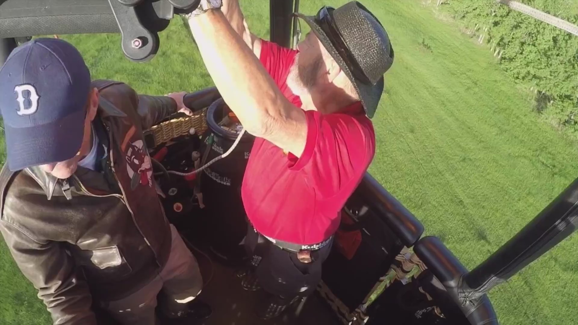 Two men in a hot air balloon basket that's in flight over a green field