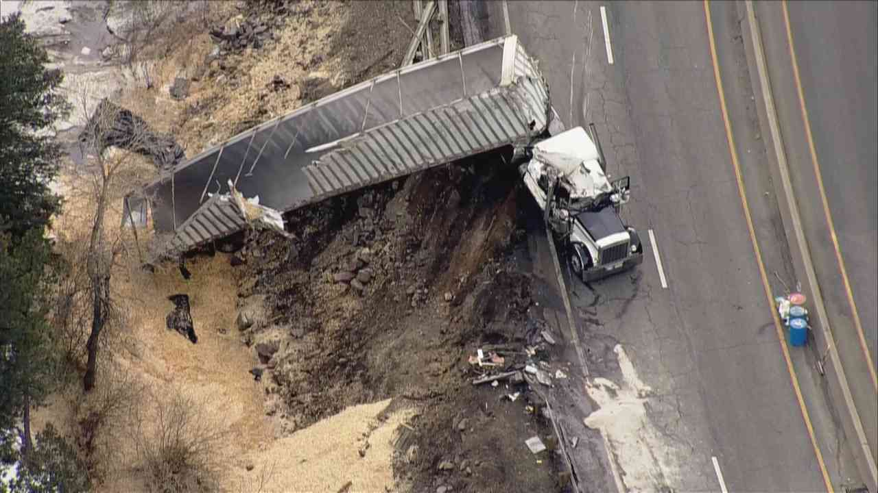 A crashed semi truck's trailer hangs over the shoulder of a highway