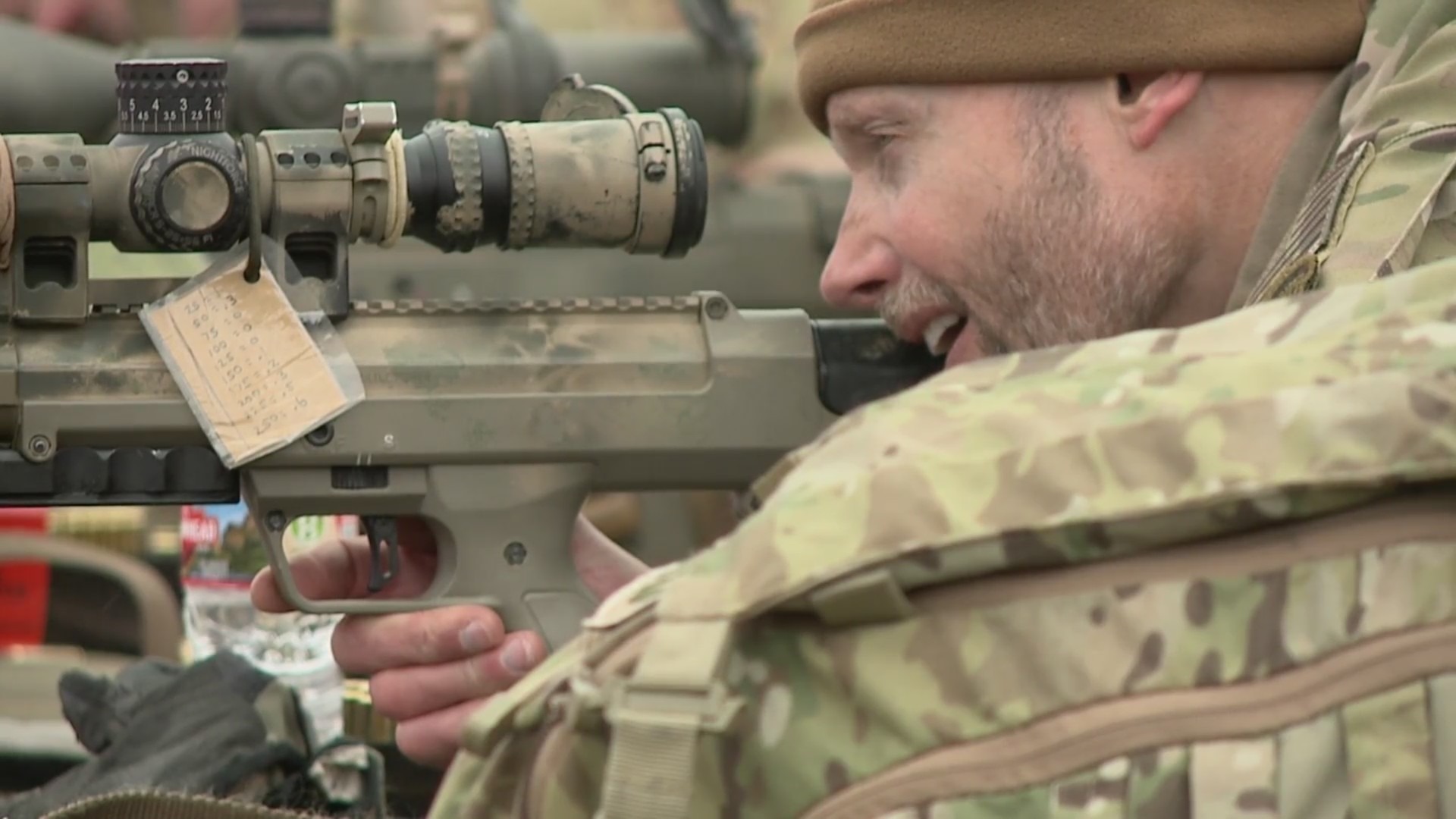 A man in desert fatigues looks into the scope of a sniper rifle