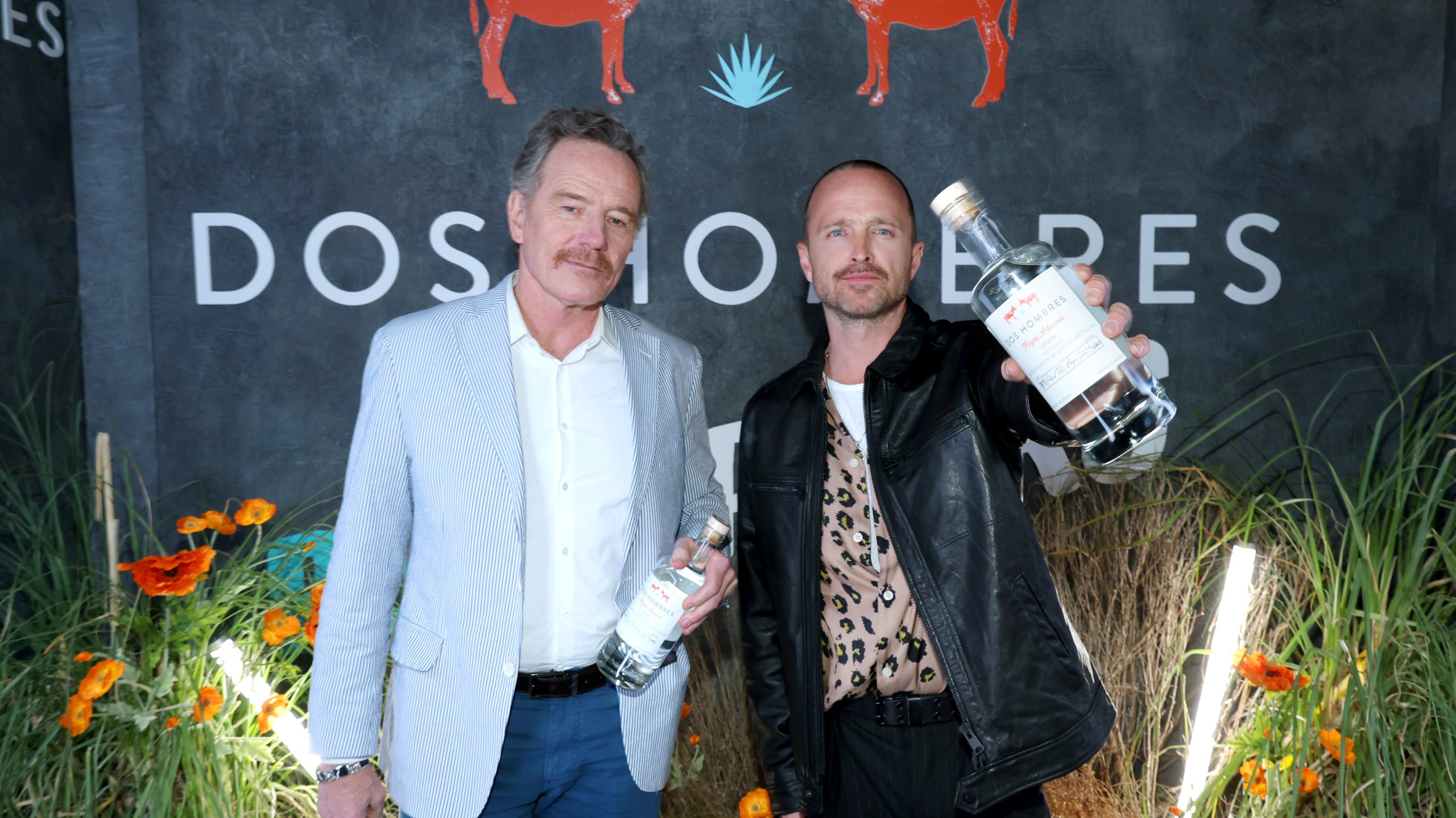 Bryan Cranston and Aaron Paul stand side by side holding bottles of mezcal