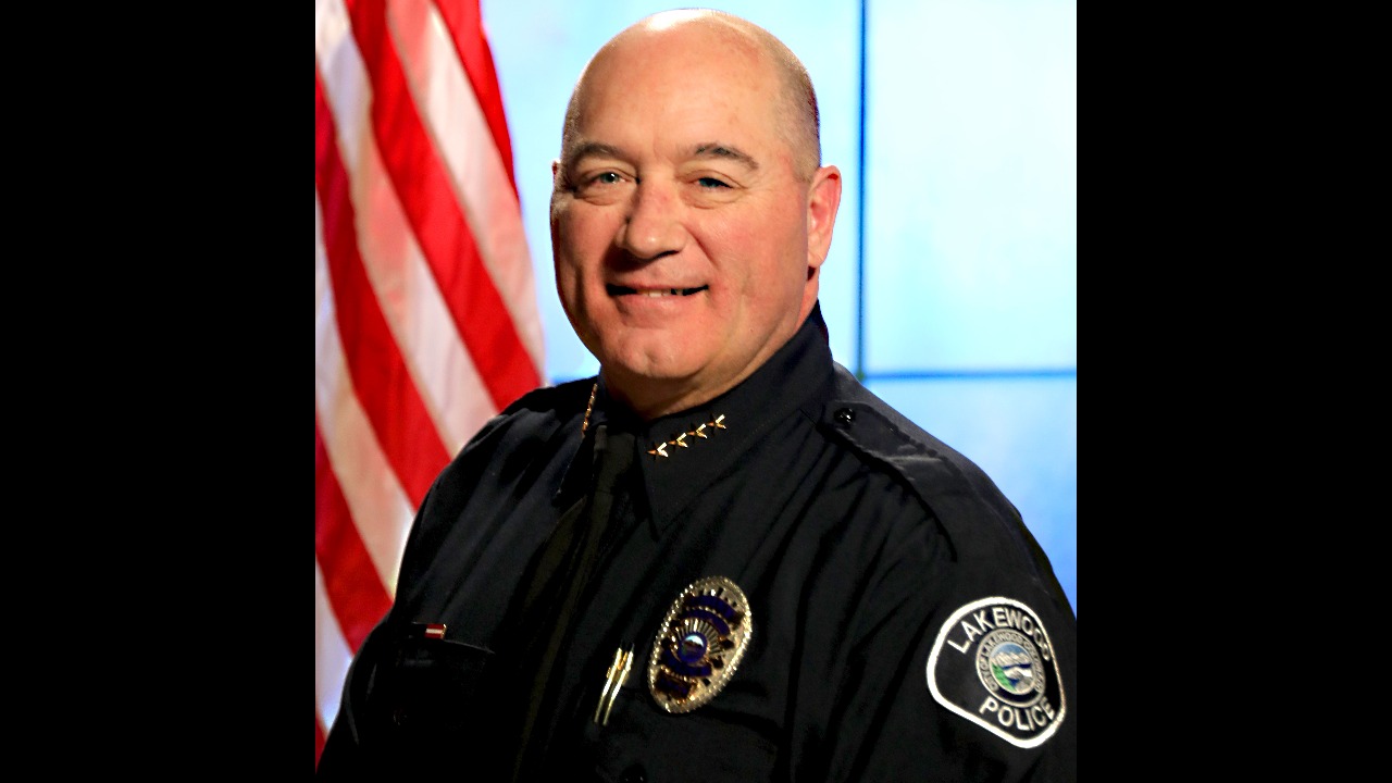 Portrait of a man in police uniform smiling at the camera in front of a U.S. flag