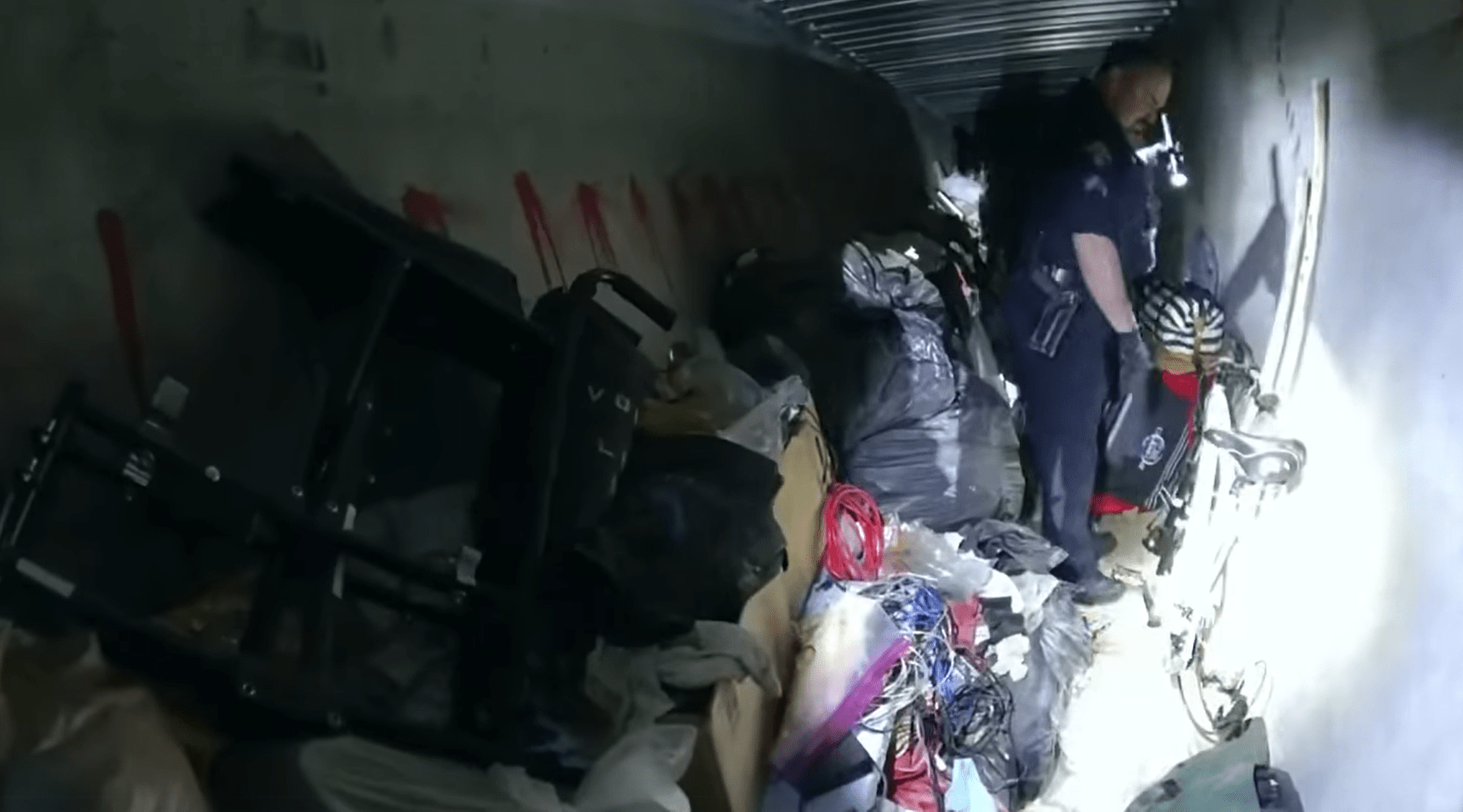 A uniformed officer inside a dark tunnel, surrounded by clutter, shines a flashlight