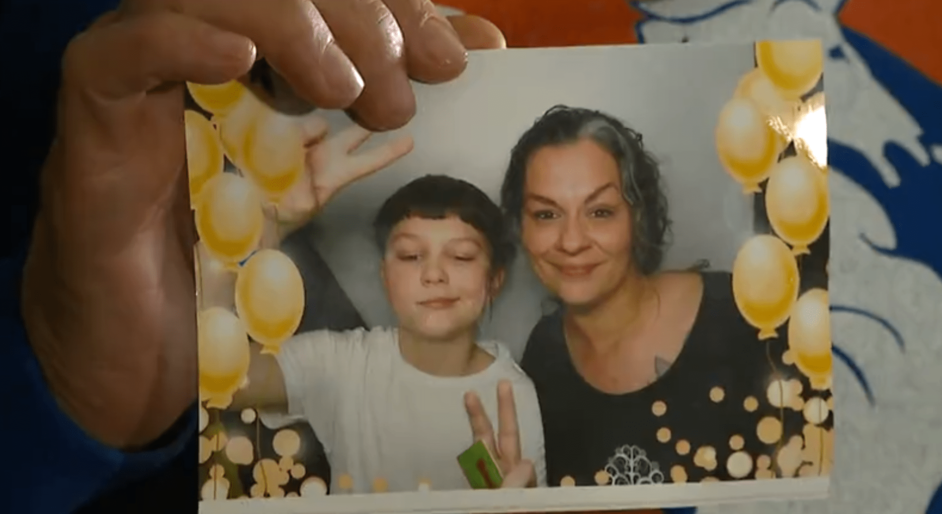 A hand holds up a picture of a boy holding up peace signs next to his mother, both smiling