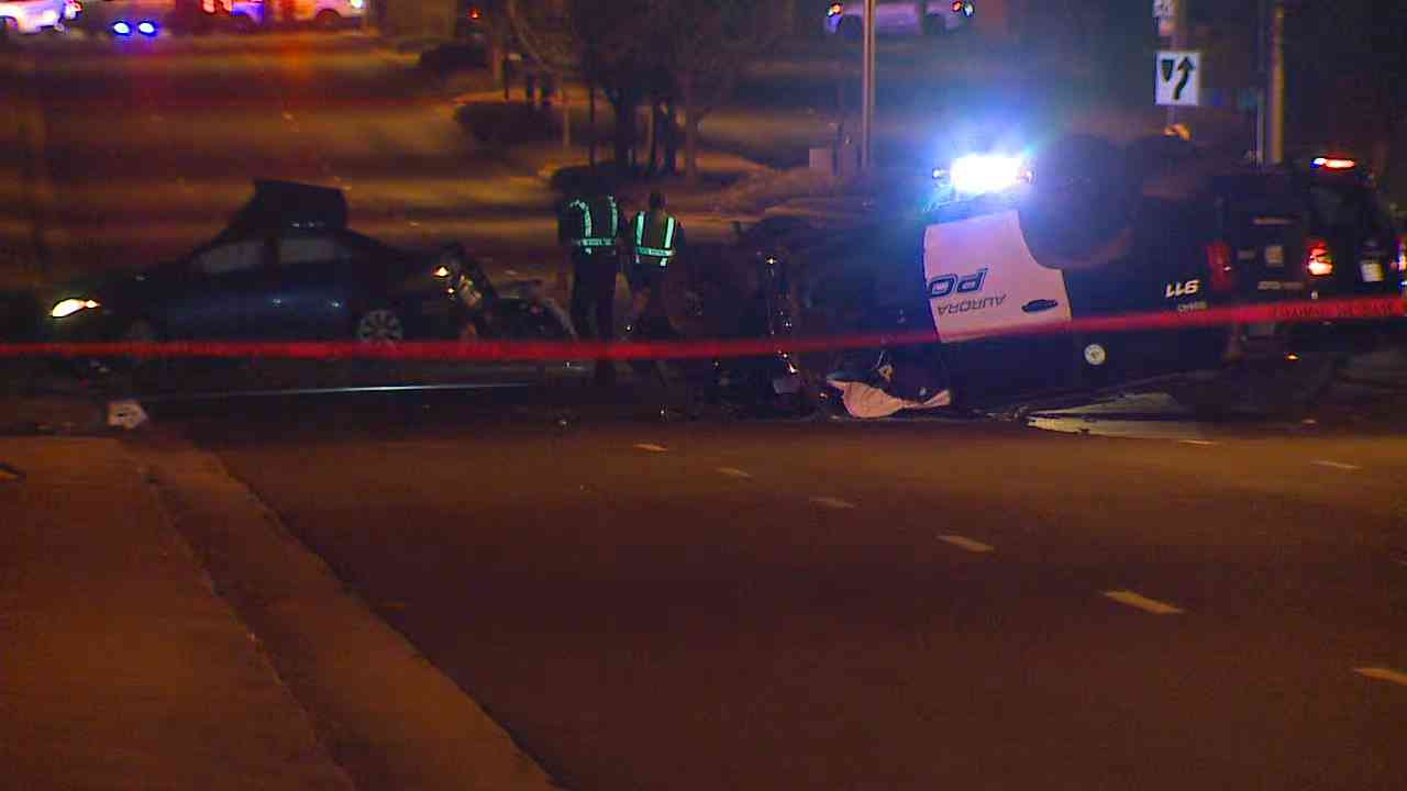 Police tape at the foreground of a crash scene showing a flipped Aurora police SUV and a sedan