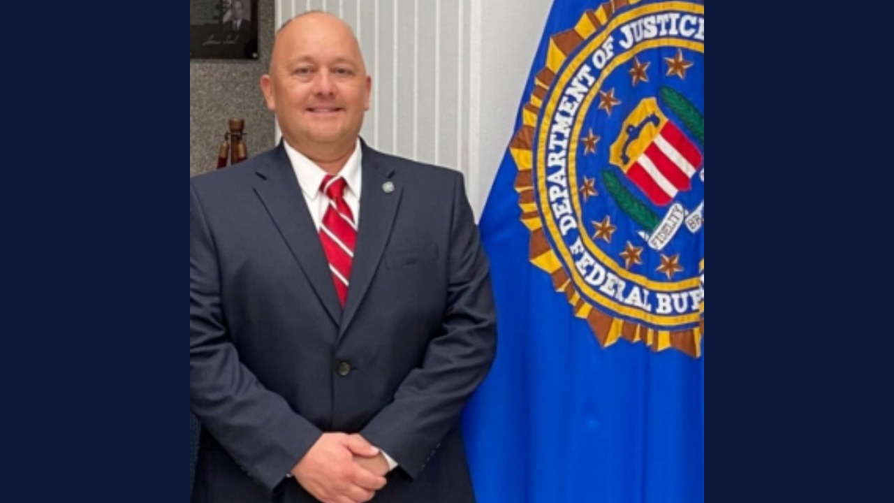 A man in a suit and red tie stands smiling at the camera with his hands folded while standing next to an FBI flag