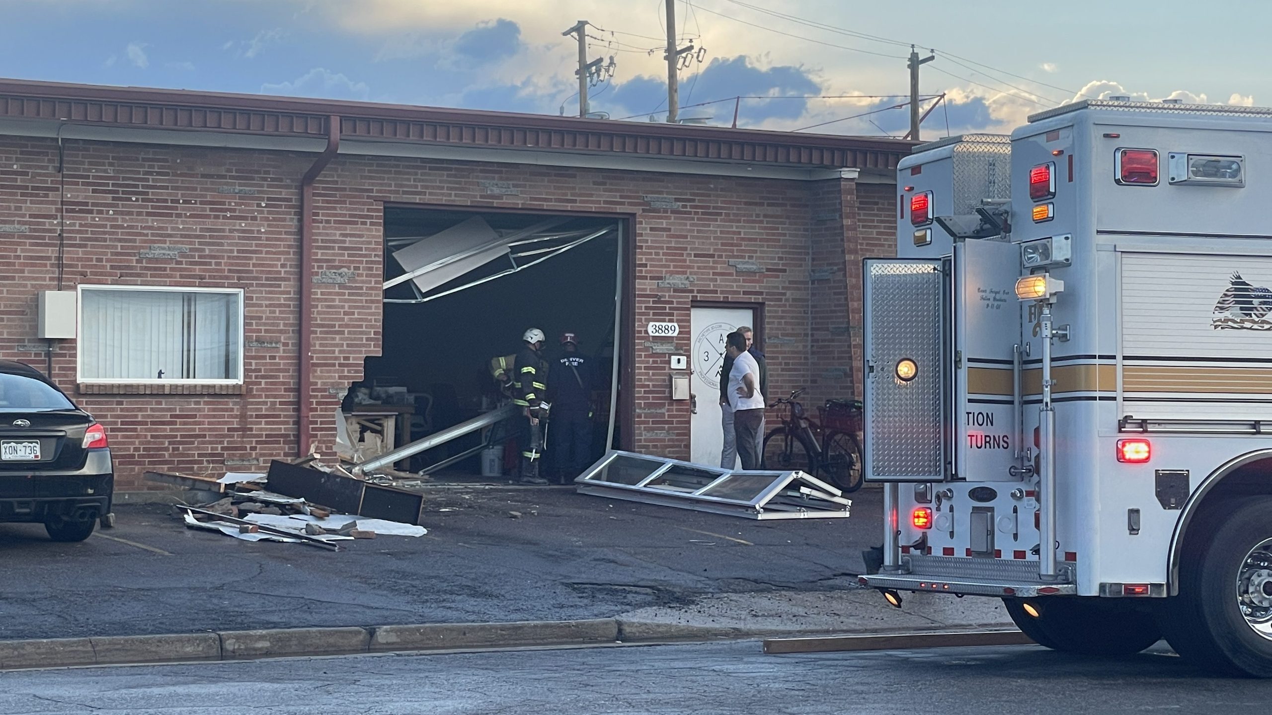 A brick building with a knocked out garage door and missing bricks, with fire personnel and truck on the scene