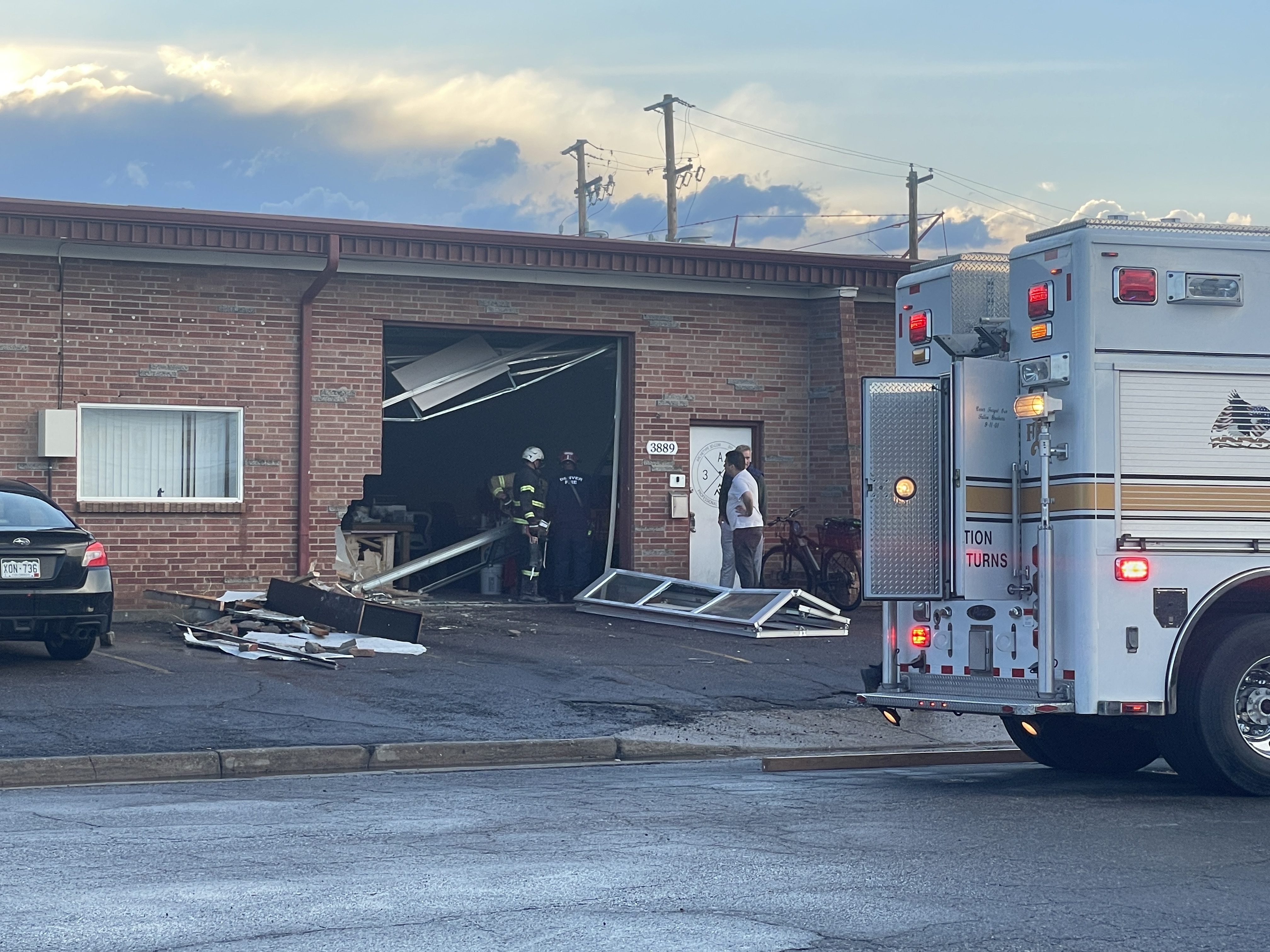 A brick building with a knocked out garage door and missing bricks, with fire personnel and truck on the scene