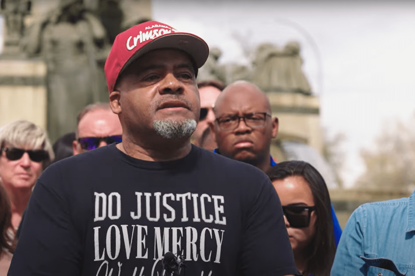 A man in a baseball cap with a T-shirt that says "DO JUSTICE LOVE MERCY" stands before a crowd