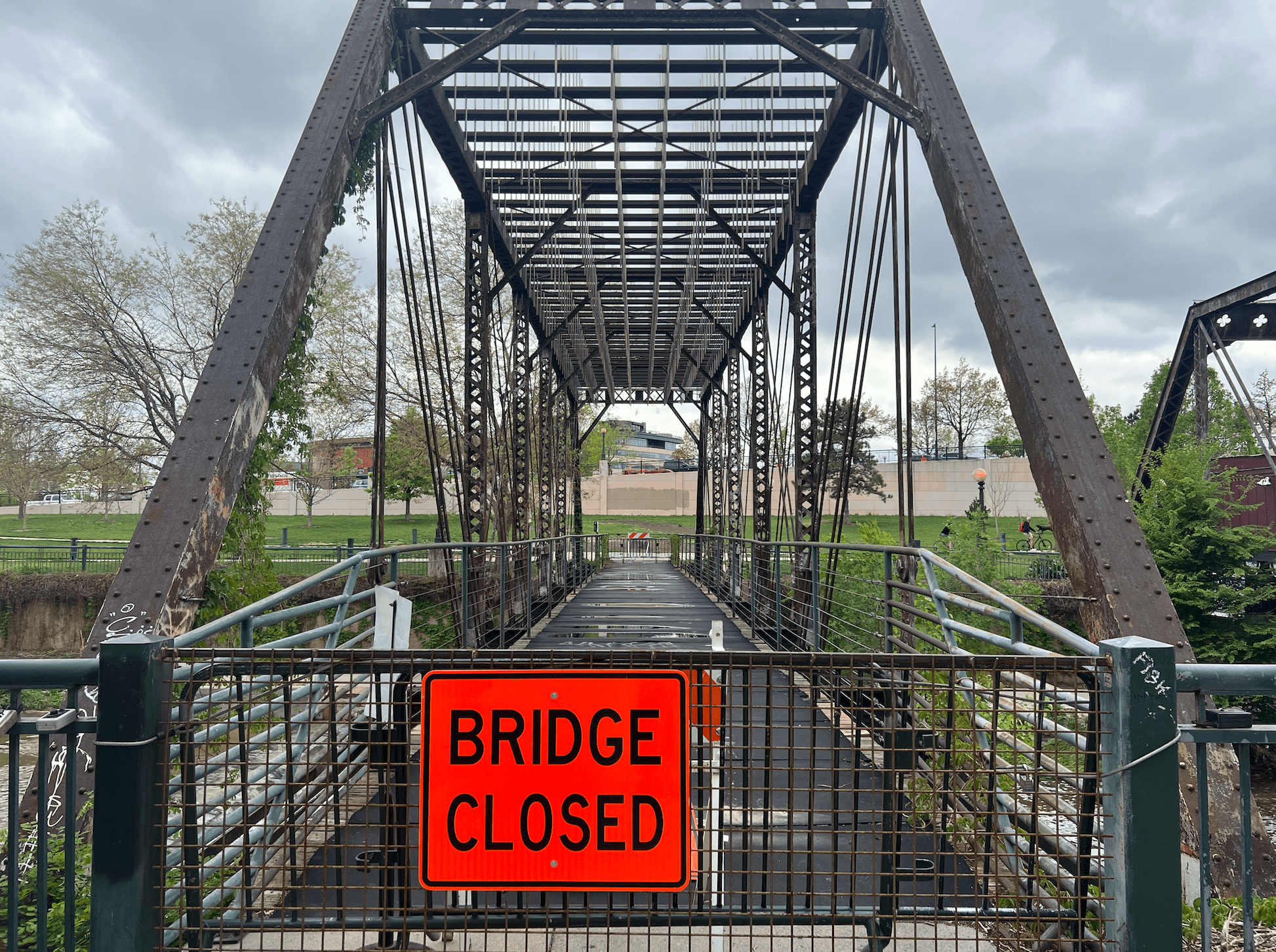 A fluorescent orange road sign says "BRIDGE CLOSED" is posted to a pedestrian bridge crossing a waterway