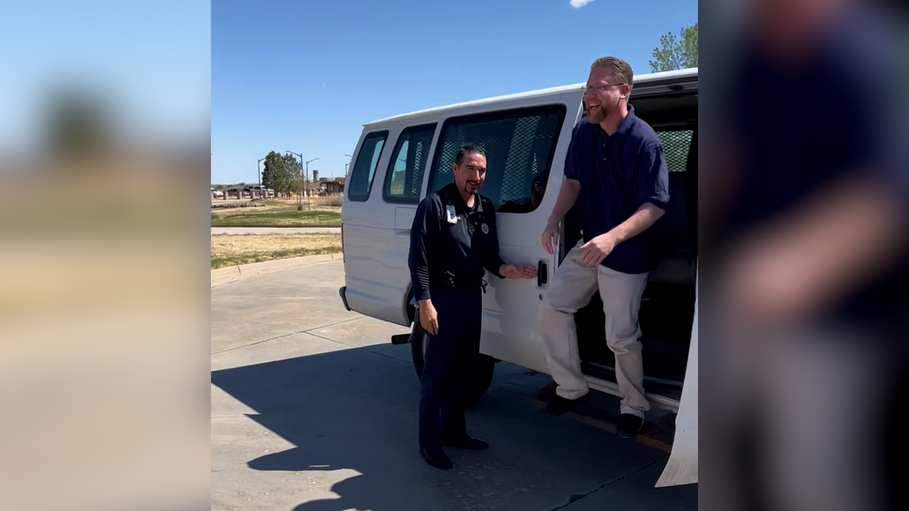 A man smiles as he gets out of a van with a guard standing by